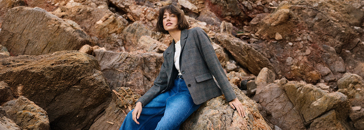 Femme portant un veston gris avec blouse blanche et jeans bleu au milieu d’un décor rocheux, look de la collection Part Two.