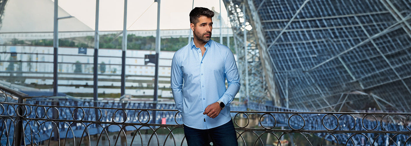 Modèle homme portant une surchemise bleu clair dans un décor urbain moderne avec structures métalliques. Styles intemporels Au Noir disponibles toute l'année avec coupes élégantes et design signature.