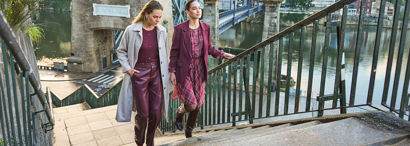 Deux femmes en looks automne, avec manteau gris, pantalon bordeaux, veston bordeaux et robe à carreaux, marchant près d’un canal urbain.