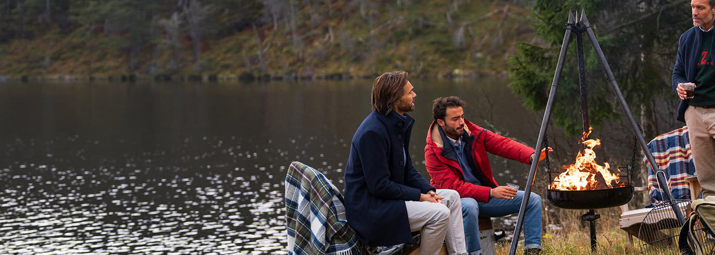 Trois hommes en vêtements automne hiver près d’un feu au bord d’un lac, portant manteaux, veste rouge, tricot et pantalon décontracté.