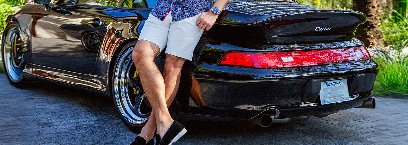 Modèle homme assis sur une voiture sportive noire portant un bermuda clair et chemise à motif. Bermudas Au Noir stylés et confortables parfaits pour la saison estivale.