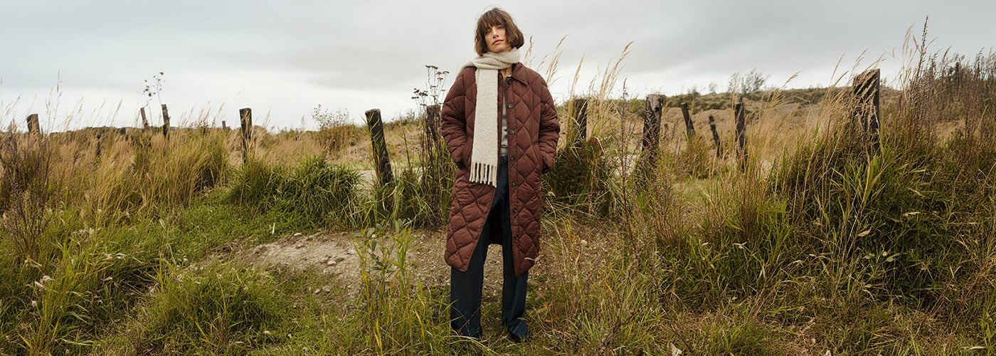 Long manteau matelassé brun accompagné d’une écharpe crème et d’un pantalon foncé, look automnal pour femme en pleine nature.