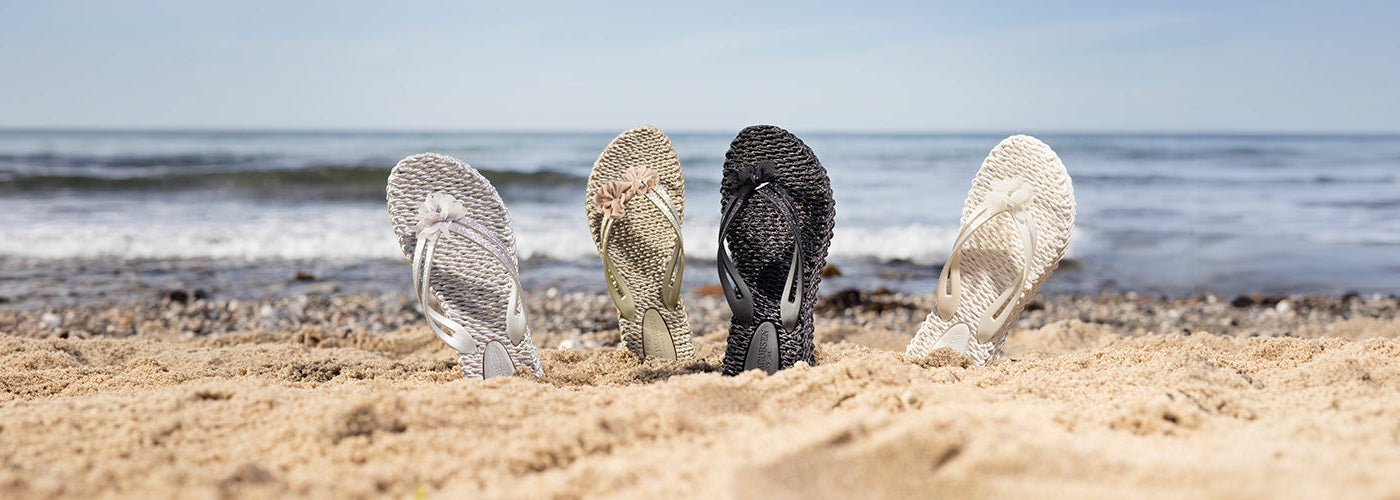Sandales tressées pour femmes disposées dans le sable sur une plage avec l’océan en arrière plan. Collection chaussures femmes été, sandales élégantes et confortables.