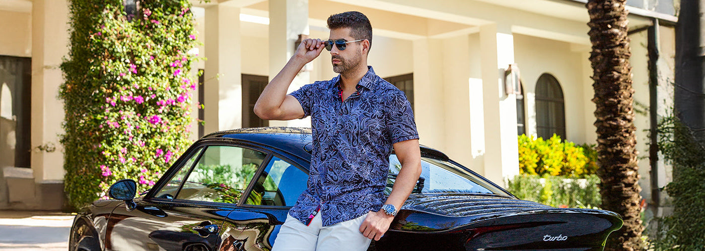 Modèle homme portant une chemise M.C. à motif tropical devant une résidence luxueuse entourée de palmiers. Chemises Au Noir légères et stylées idéales pour un look estival sophistiqué.