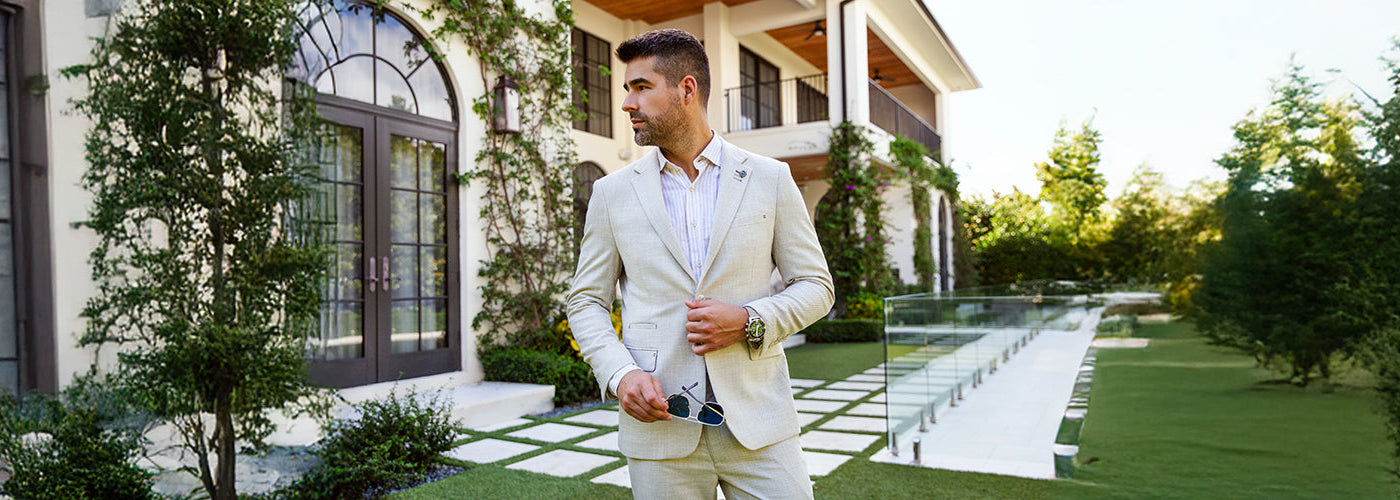 Modèle homme élégant portant un complet beige devant une résidence luxueuse entourée de jardins modernes. Habits Au Noir sophistiqués pour un style distingué et contemporain.