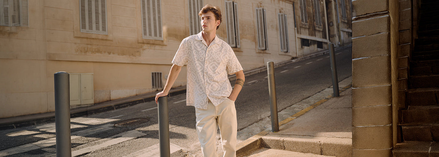 Homme portant une chemise blanche à motifs ton sur ton et un pantalon clair dans une rue ensoleillée, pour un look Garcia homme estival, urbain et casual chic
