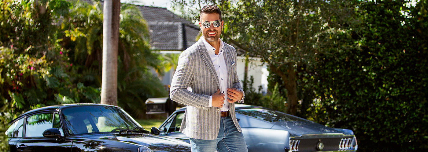 Modèle homme élégant portant un Blazer rayé devant une voiture sportive noire dans un décor tropical. Collection complète Au Noir avec styles modernes, vêtements premium et looks raffinés pour hommes.