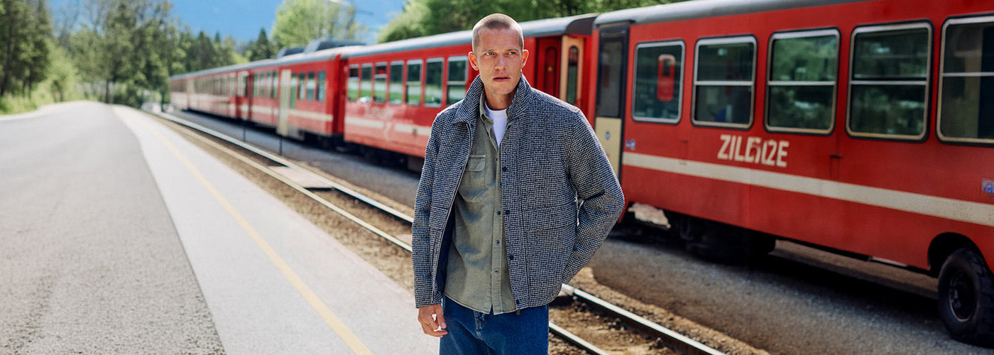 Homme portant une veste texturée grise, chemise kaki et jeans devant un train rouge, look masculin de la collection Matinique.