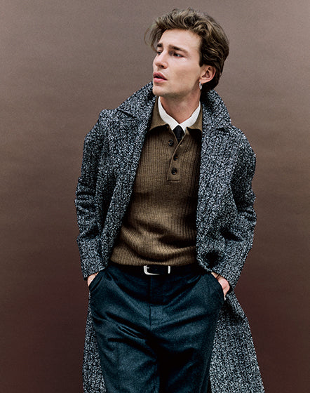Homme portant un long manteau texturé gris sur un tricot brun à col boutonné et un pantalon foncé, look homme automne hiver.
