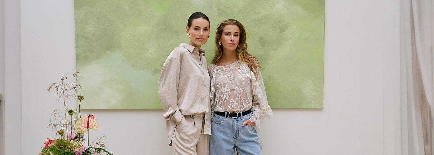 Deux modèles femmes portant des hauts élégants dans un décor intérieur lumineux avec tableau vert et plantes. Collection hauts femmes mode, chemisiers et chandails tendance.