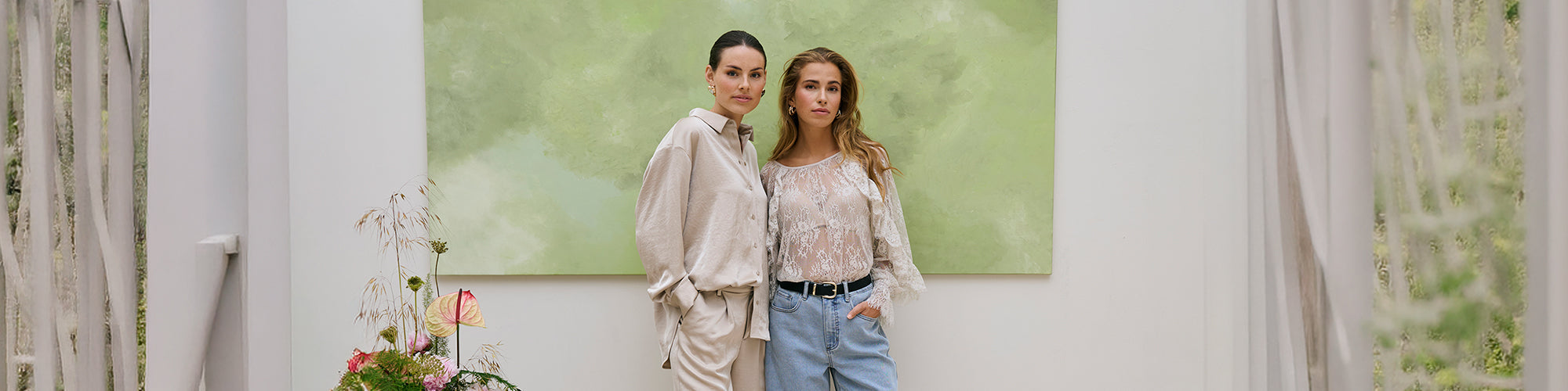 Deux modèles femmes portant des hauts élégants dans un décor intérieur lumineux avec tableau vert et plantes. Collection hauts femmes mode, chemisiers et chandails tendance.