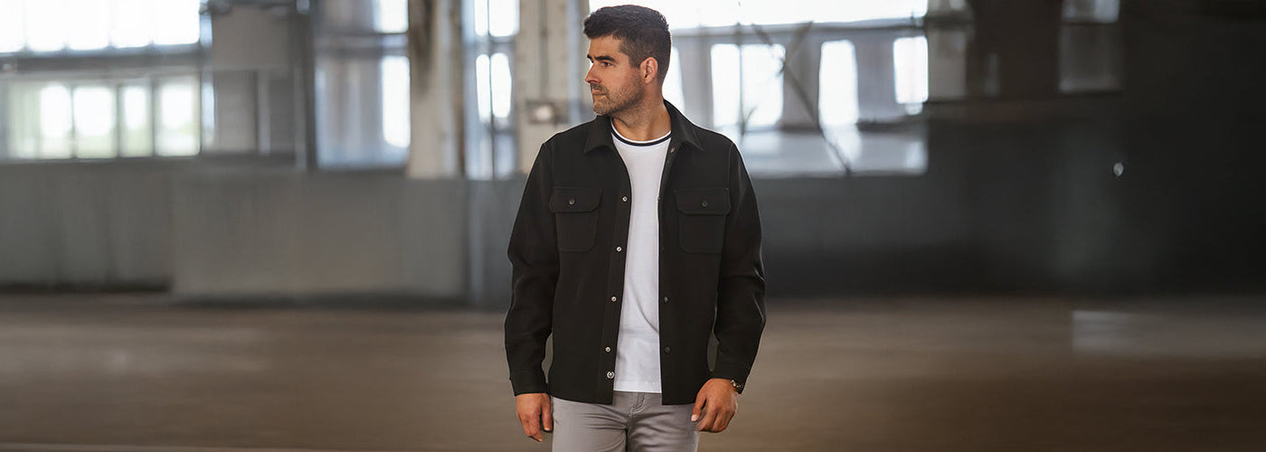 Homme en surchemise noire et pantalon gris, dans un vaste espace intérieur lumineux, regardant vers la gauche.
