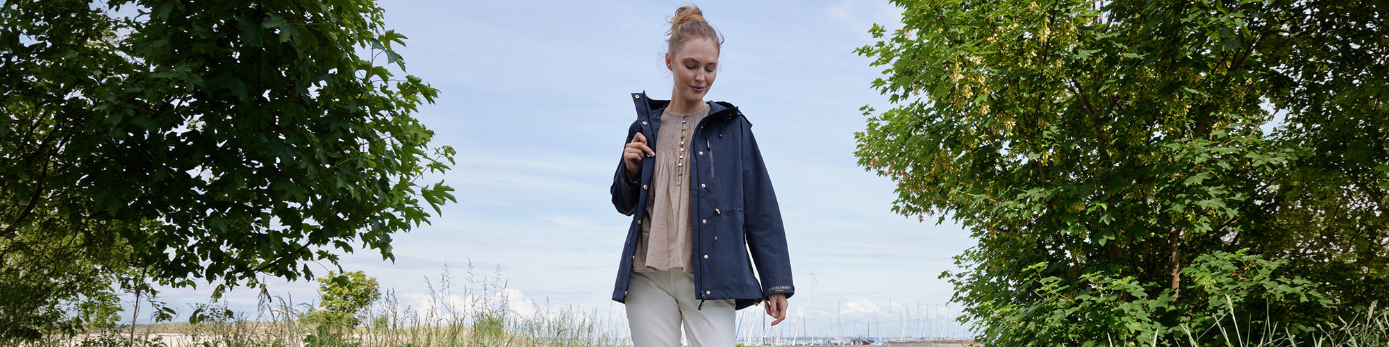 Modèle femme portant un coupe vent bleu dans un décor naturel avec arbres et port en arrière plan. Collection coupe vent femmes, manteaux légers et vêtements extérieurs.