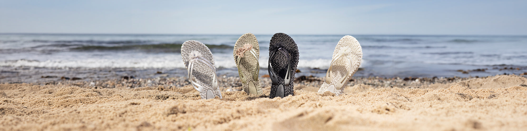 Sandales tressées pour femmes disposées dans le sable sur une plage avec l’océan en arrière plan. Collection chaussures femmes été, sandales élégantes et confortables.