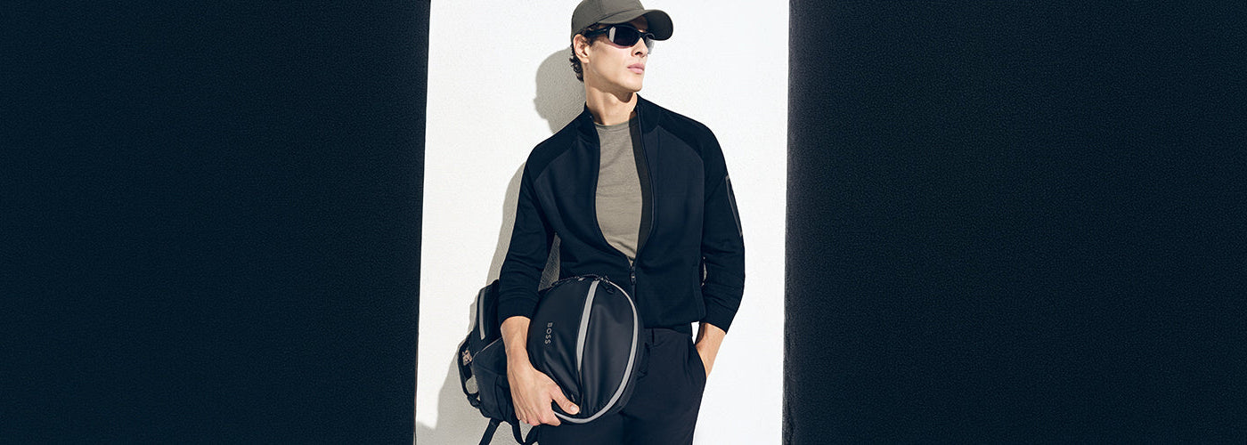 Modèle homme portant une casquette et des lunettes de soleil avec un sac de sport noir dans un décor moderne minimaliste. Collection accessoires hommes mode, sacs et accessoires élégants.
