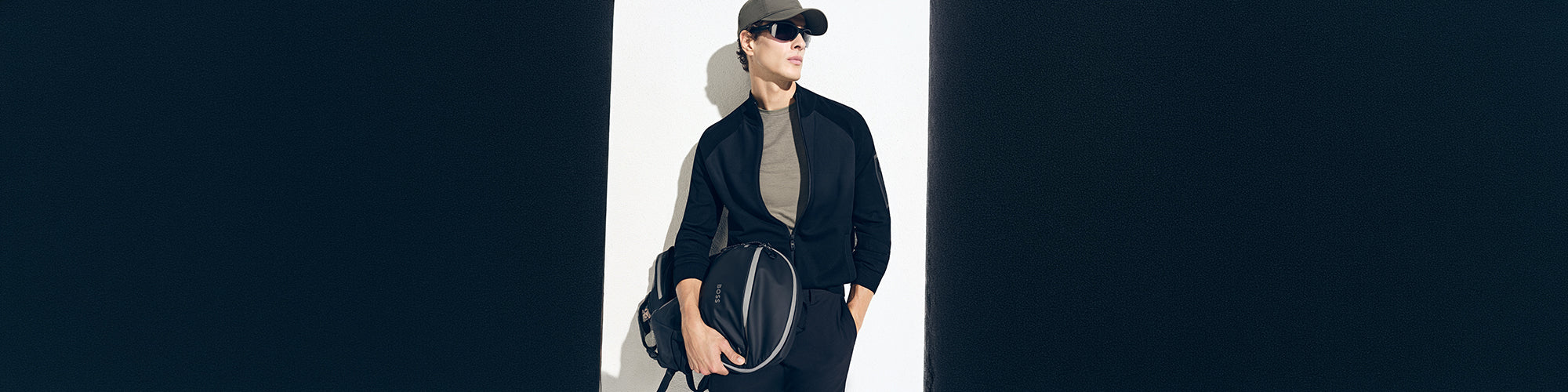 Modèle homme portant une casquette et des lunettes de soleil avec un sac de sport noir dans un décor moderne minimaliste. Collection accessoires hommes mode, sacs et accessoires élégants.
