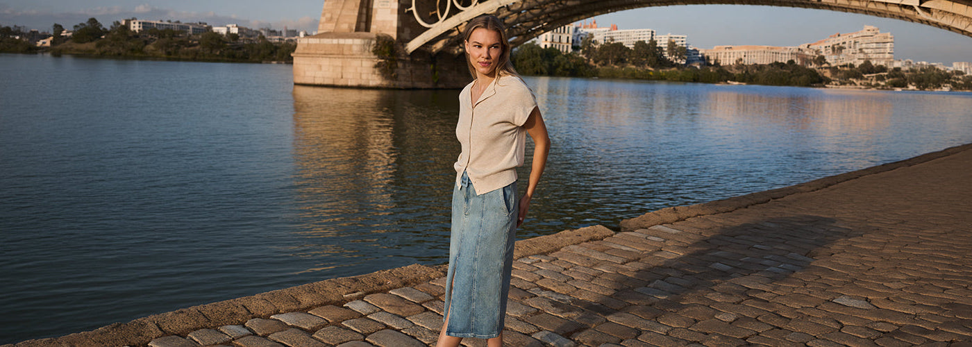 Femme portant un haut beige à manches courtes et une jupe longue en denim bleu au bord de l’eau sous un pont