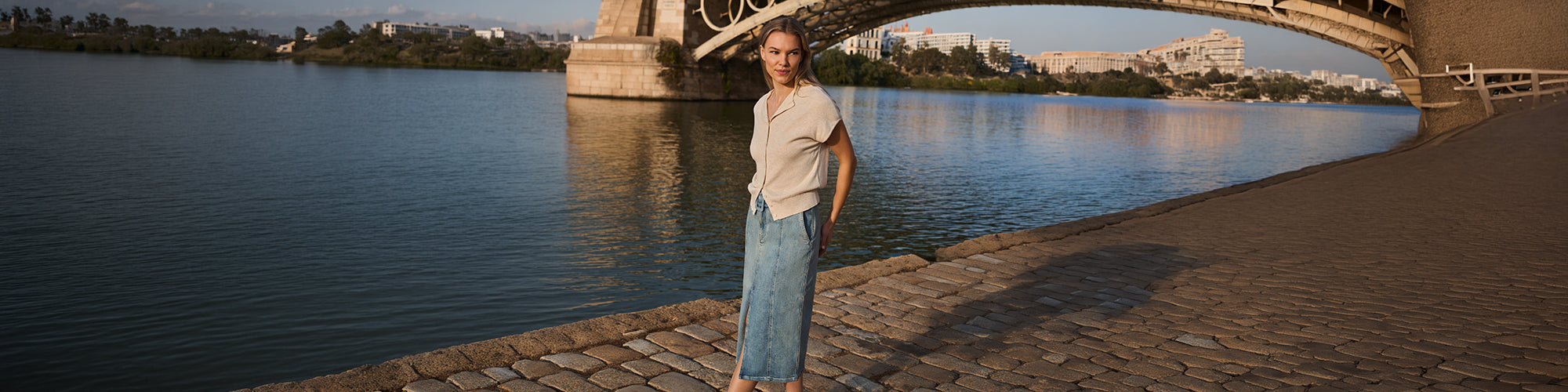 Femme portant un haut beige à manches courtes et une jupe longue en denim bleu au bord de l’eau sous un pont