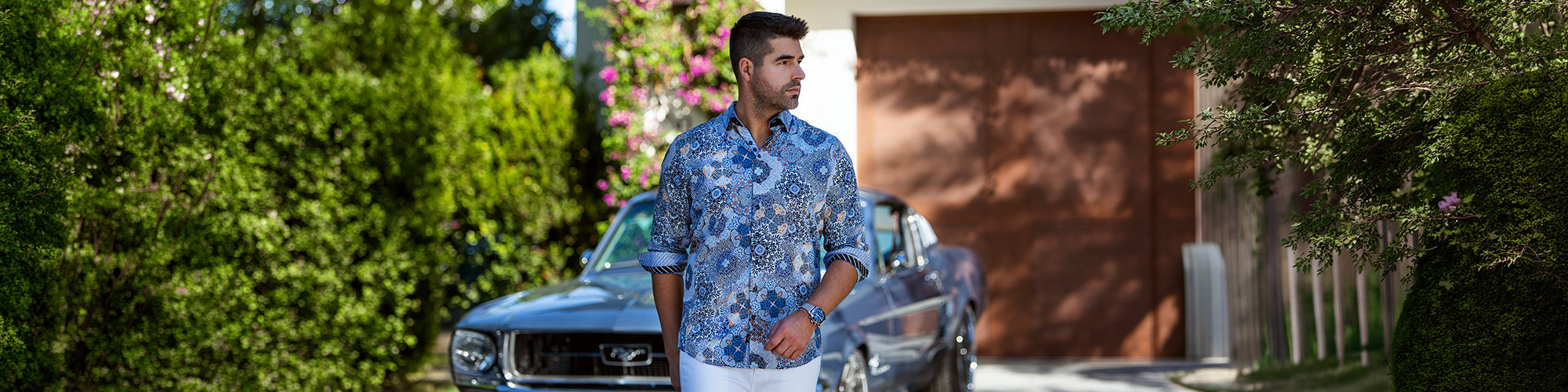 Modèle homme portant une chemise bleue à motifs devant une voiture classique dans un jardin ensoleillé. Collection chemises hommes élégantes et chemises habillées tendance.
