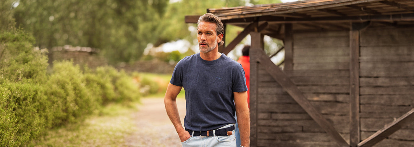 Modèle homme portant un T-Shirt bleu marine dans un décor champêtre près d’une grange en bois. Collection hauts hommes mode et T-Shirt confortables.