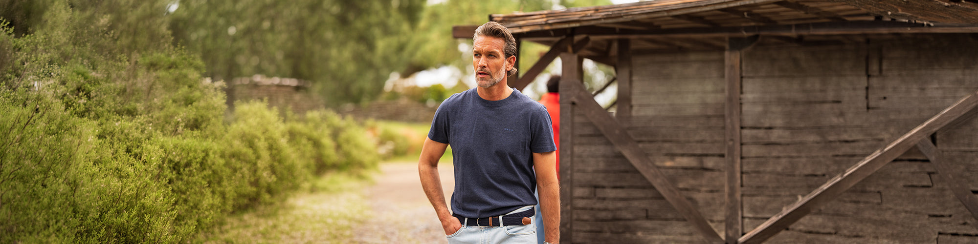 Modèle homme portant un T-Shirt bleu marine dans un décor champêtre près d’une grange en bois. Collection hauts hommes mode et T-Shirt confortables.