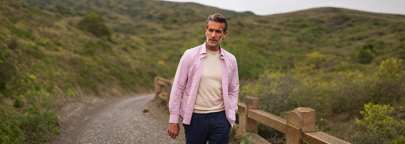 Modèle homme marchant sur un chemin naturel dans les collines portant chemise rose ouverte sur un T-Shirt beige avec pantalon foncé. Collection vêtements hommes élégants et styles modernes.