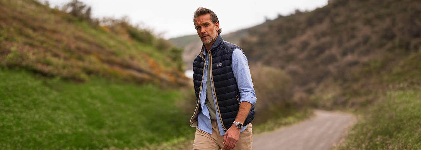 Modèle homme marchant sur un chemin de campagne portant un coupe vent sans manches matelassé avec chemise bleue. Collection coupe vent hommes et manteaux légers pour l’extérieur.