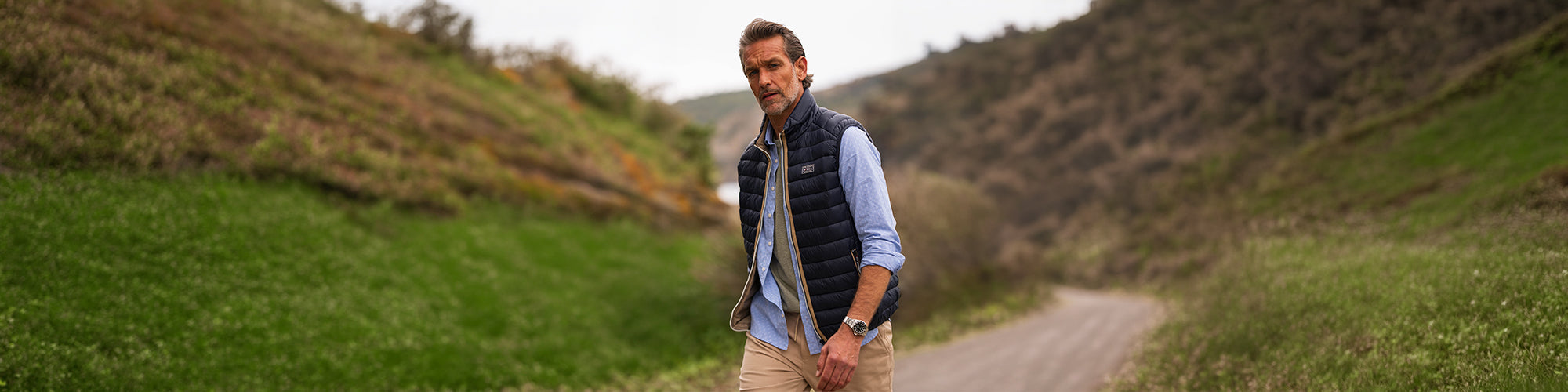Modèle homme marchant sur un chemin de campagne portant un coupe vent sans manches matelassé avec chemise bleue. Collection coupe vent hommes et manteaux légers pour l’extérieur.