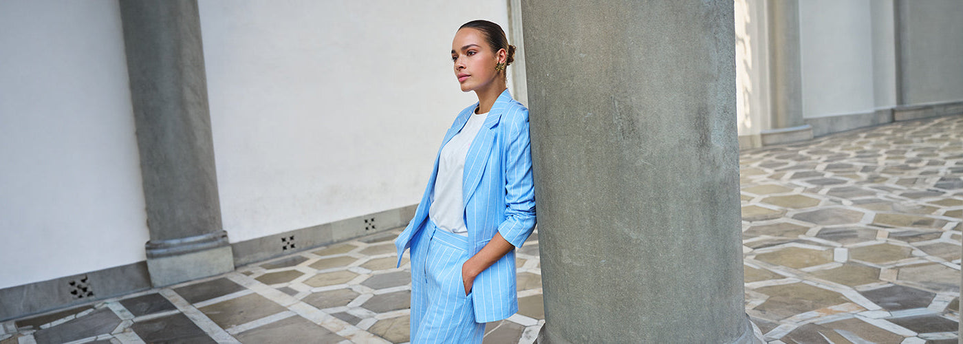 Modèle femme portant un veston bleu rayé avec pantalon assorti dans une galerie élégante avec colonnes. Collection Blazer femmes chic et tailleurs modernes.