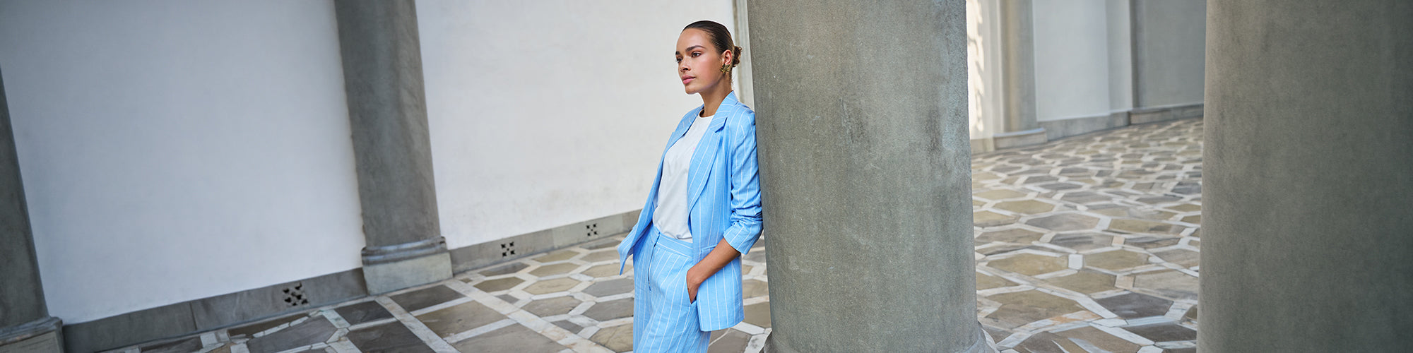 Modèle femme portant un veston bleu rayé avec pantalon assorti dans une galerie élégante avec colonnes. Collection Blazer femmes chic et tailleurs modernes.