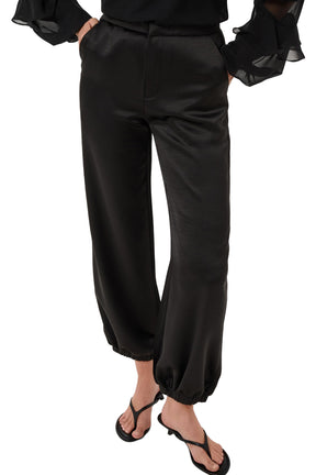 Cream women's close front view of black satin-look jogger pants showing waistband, fly seam, and elastic hems