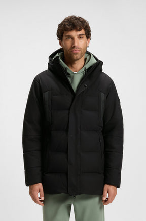 Vue avant sur modèle du manteau d’hiver noir à capuchon avec rabat et poches zippées pour homme par HUGO BOSS