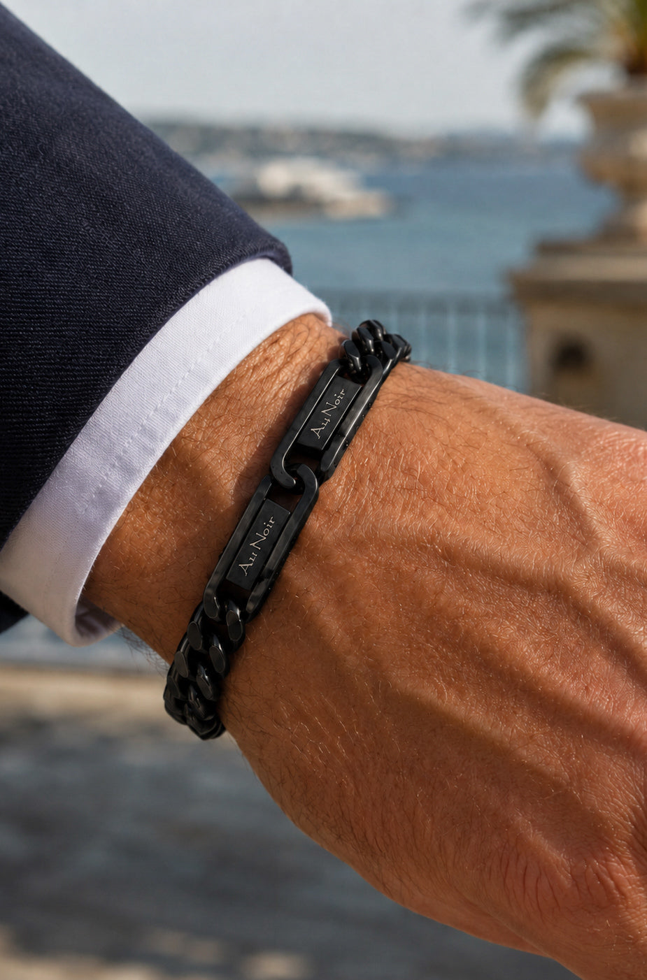 Détail: Bracelet chaîne noir porté au poignet avec plaques gravées Au Noir près d’un poignet de chemise pour homme