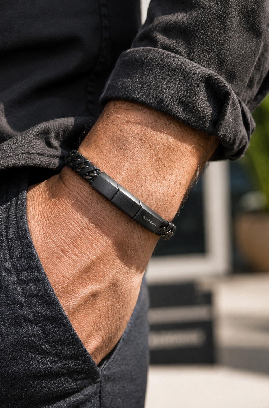 Détail: Bracelet noir tressé porté au poignet avec fini foncé et fermoir rectangulaire à logo pour homme par Au Noir