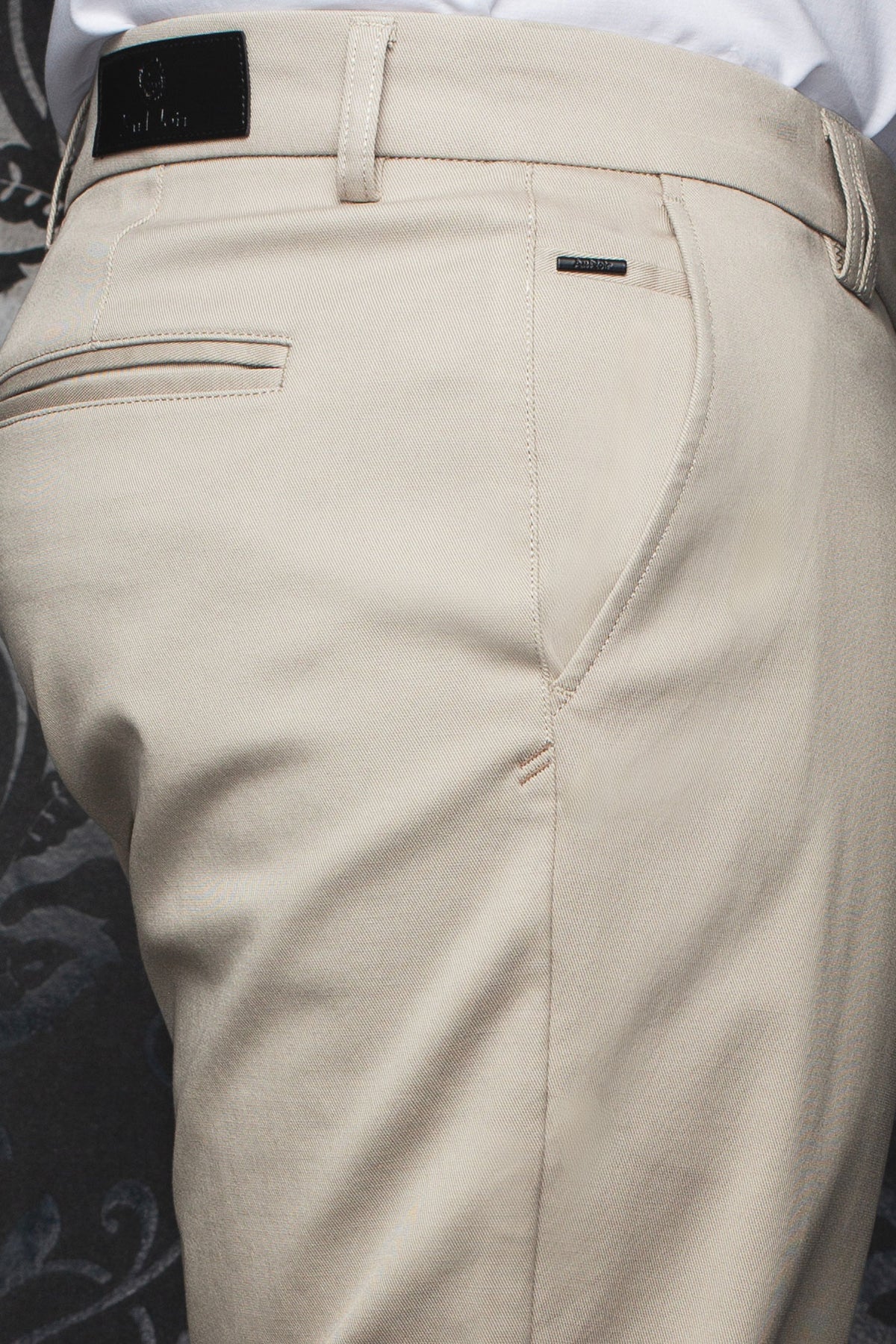 Détail rapproché du pantalon chino beige avec écusson noir, passants et poche passepoilée pour homme par Au Noir