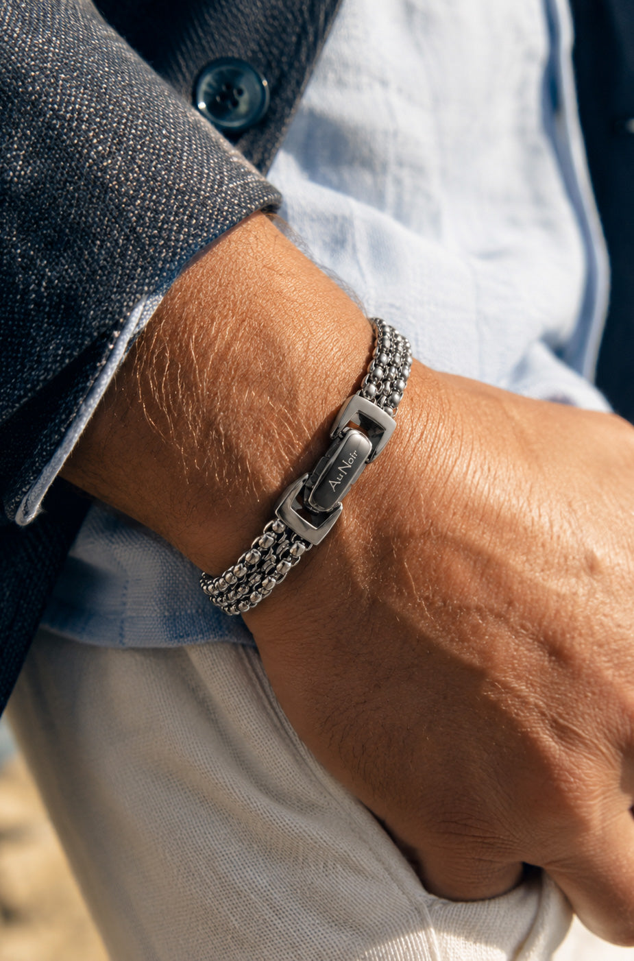 Détail: Bracelet argenté à maillons multiples porté au poignet, fermoir à logo visible pour homme par Au Noir