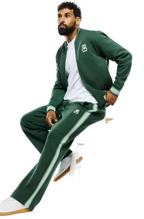 Men's angled seated view of dark green sweatpants showing the light green side stripe and thigh logo patch by Psycho Bunny