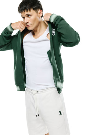 Psycho Bunny men's angled model view of open dark green bomber jacket showing zipper, pocket and striped cuffs, side view