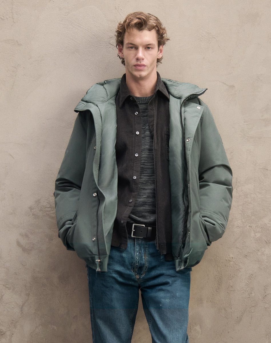 Manteau vert superposé à une chemise foncée et un tricot gris, complété par un jeans bleu pour une tenue automnale homme.
