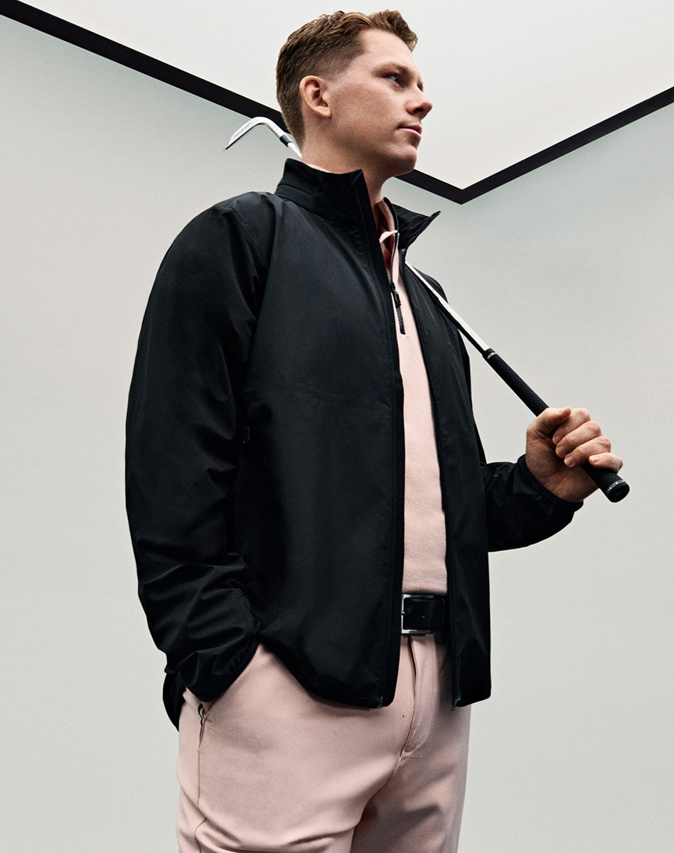 Veste BOSS noire avec polo et pantalon rose pâle, tenue de golf pour homme complétée par un bâton.
