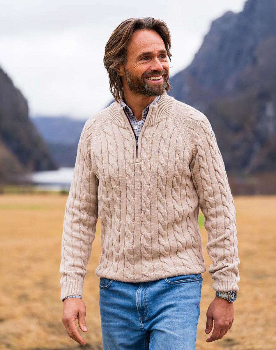 Beige cable knit sweater with zip collar and blue jeans, warm men’s look in a natural setting.
