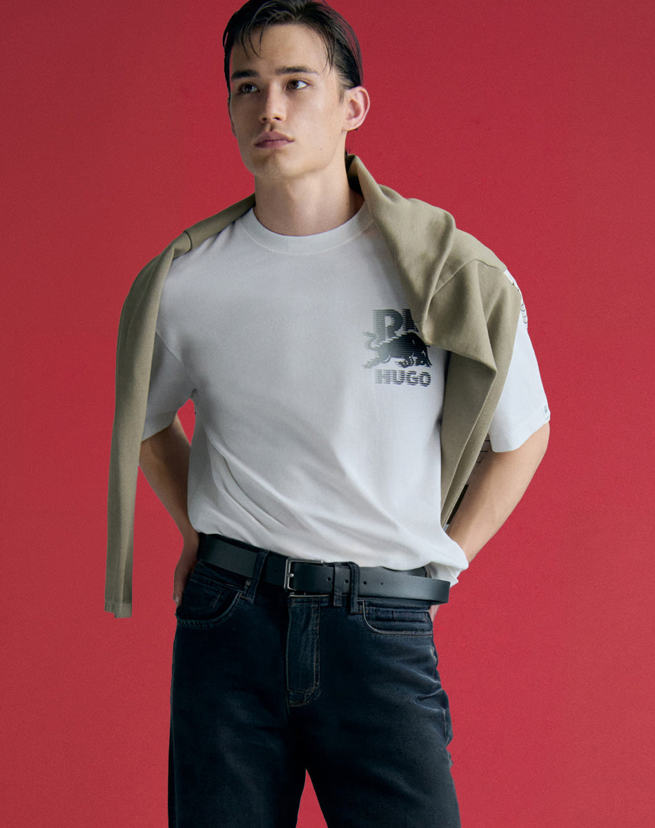 T-Shirt HUGO blanc à motif graphique porté avec jeans noir et chandail beige posé sur les épaules, style urbain pour homme.
