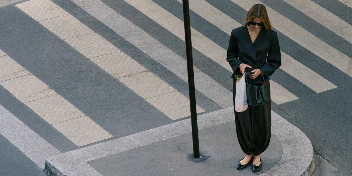 Look InWear noir composé d’un veston cintré et d’un pantalon ample, accompagné de ballerines et d’un sac dans un décor urbain.

